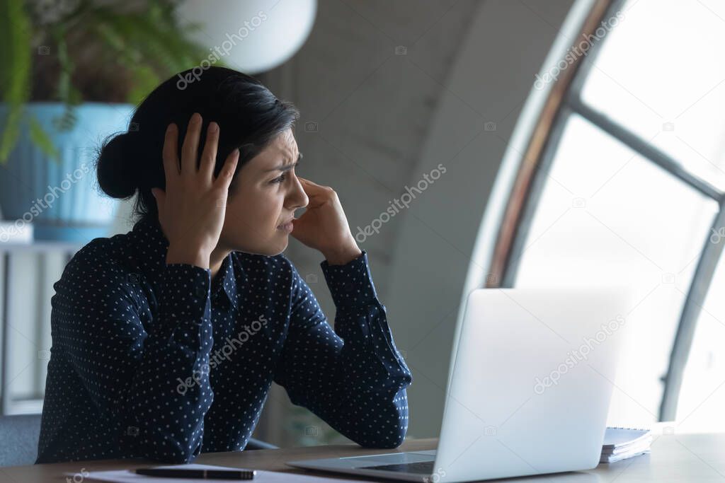 Overworked Indian employee sitting at workplace suffering from headache