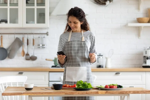 Happy young attractive woman cooking to recipe on smartphone. — Stock Photo, Image