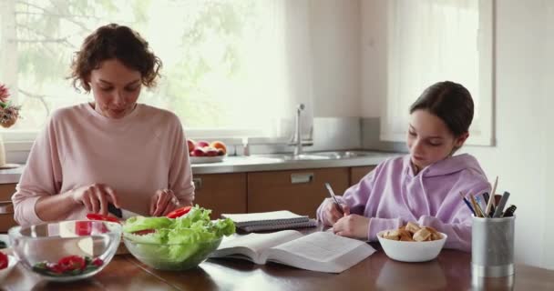 Mãe preparando salada enquanto a filha adolescente estuda na cozinha . — Vídeo de Stock