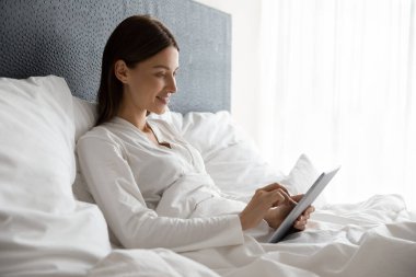 Gülümseyen kadın yatakta tablet kullanarak rahatlıyor.