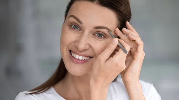 Young woman look in mirror pinch eyebrows with tweezers