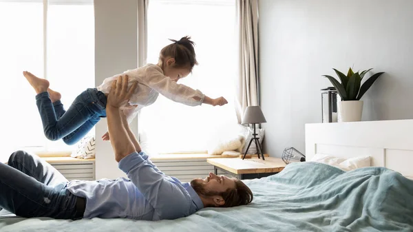 Smiling father holding little girl pretending flying, relaxing in bed