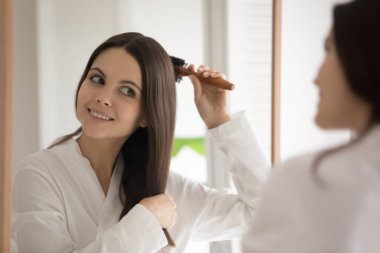 Ayna yansıması gülümseyen genç kadın saçlarını ahşap fırçayla tarıyor.