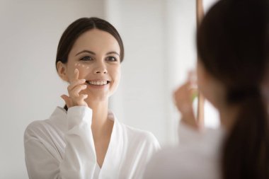 Ayna yansıması gülümseyen genç kadın nemlendirici yüz kremi sürüyor.