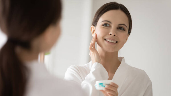 Mirror reflection smiling girl with healthy skin applying face cream