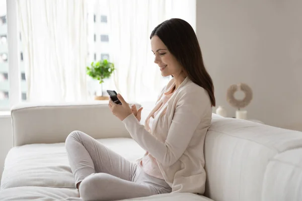 Sorridente bella donna in possesso di smartphone, seduto su un accogliente divano — Foto Stock