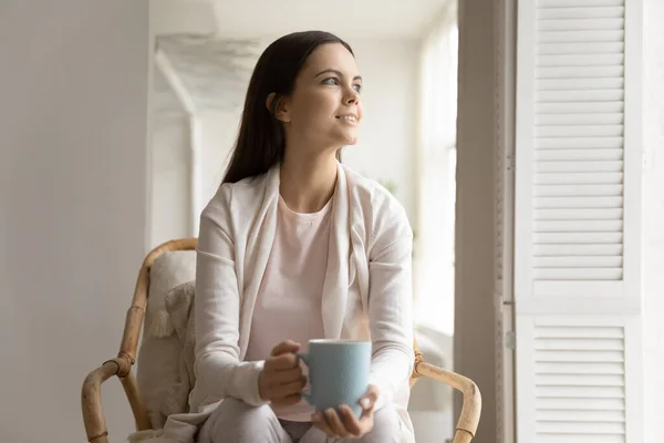 Smiling dreamy young woman holding cup, sitting in cozy armchair — Stock Photo, Image