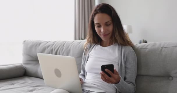 Mujer joven feliz usando el ordenador portátil y el teléfono sentado en el sofá — Vídeo de stock