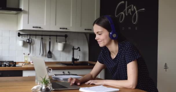 Estudiante chica hacer asignación en la cocina desgaste auriculares uso de ordenador portátil — Vídeo de stock