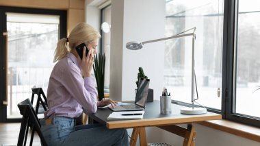 Genç beyaz kadın çalışan cep telefonuyla dizüstü bilgisayar kullanıyor.