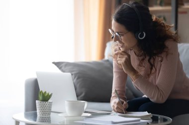 Kulaklıklı gülümseyen genç kadın dizüstü bilgisayarda ders çalışırken notlar alıyor.
