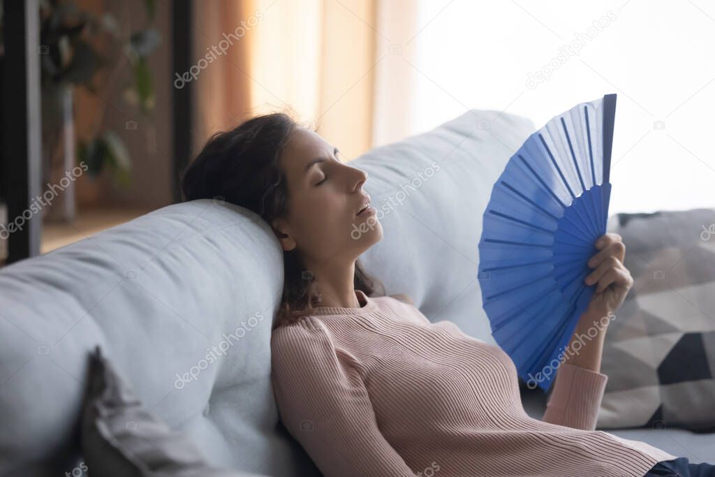 Overheated young woman waving with hand fan at home