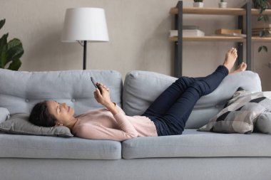 Genç beyaz kadın kanepeye uzanıp akıllı telefon kullanıyor.
