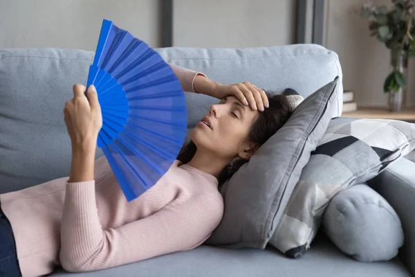 Mujer joven sobrecalentada relajarse en el sofá onda con ventilador de mano — Foto de Stock