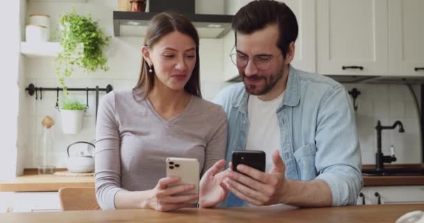 Веселая пара сидит дома на кухне и веселится со смартфонами — стоковое видео