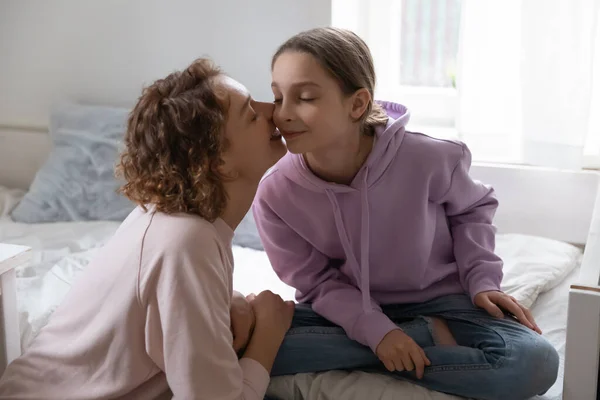 Loving mother kissing adorable teenage pretty daughter on cheek