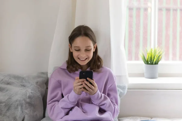 Ragazza sorridente adolescente guardando lo schermo del telefono, seduto sul letto — Foto Stock