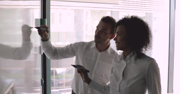 Dos colegas diversos sosteniendo marcadores dibujando en pizarra blanca en la oficina — Vídeo de stock