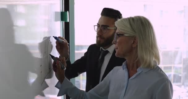 Sorridente grafico maturo mentore disegno sulla lavagna bianca con dipendente arabo — Video Stock
