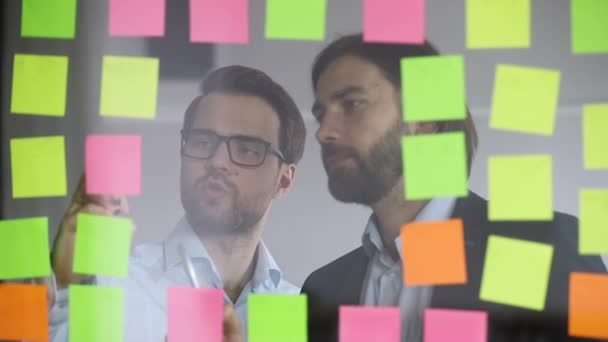 Два чоловіки-колеги пишуть на липких нотах на скляній стіні — стокове відео