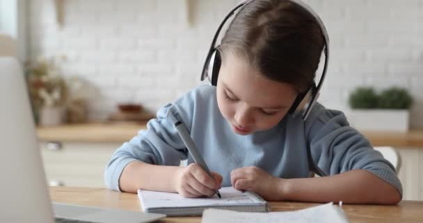 Маленька дівчинка початкової школи в навушниках, роблячи домашнє завдання віддалено . — стокове відео