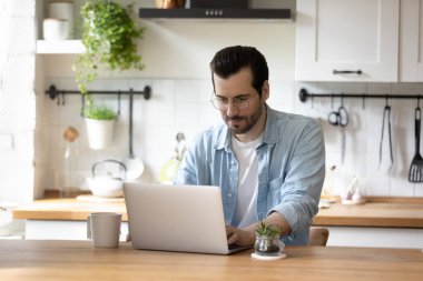 Beyaz adam mutfakta oturup dizüstü bilgisayarla çalışıyor.
