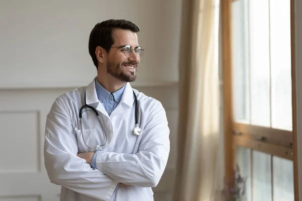 Smiling young doctor planning future career, standing near window. — Stock Photo, Image