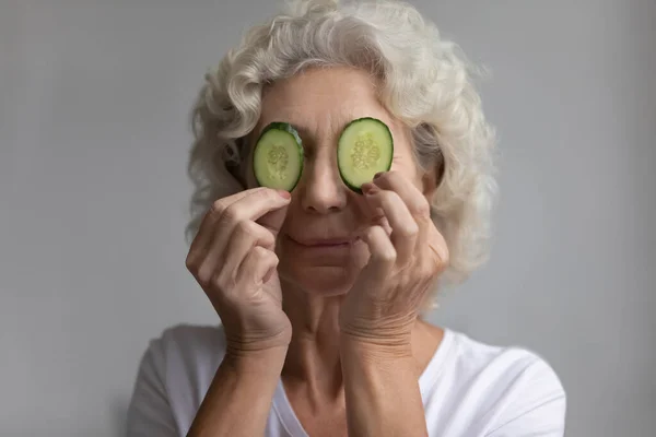 Elderly woman covers eyes with cucumbers slices enjoy skincare procedure — Stock Photo, Image