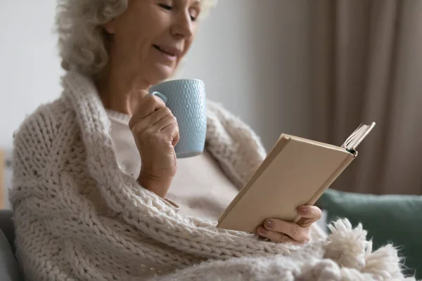 Gammal kvinna koppla av på soffan dricka te läsa en bok — Stockfoto