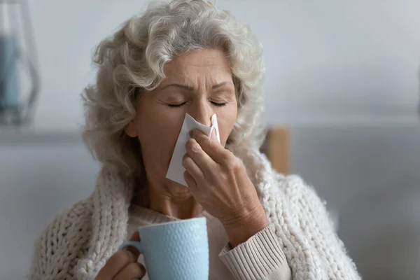 Unhealthy elderly woman blowing runny nose drinks antiviral beverage — Stock Photo, Image