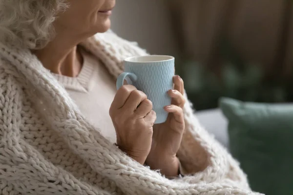 Aged calm woman wrapped in knitted plaid drink tea closeup