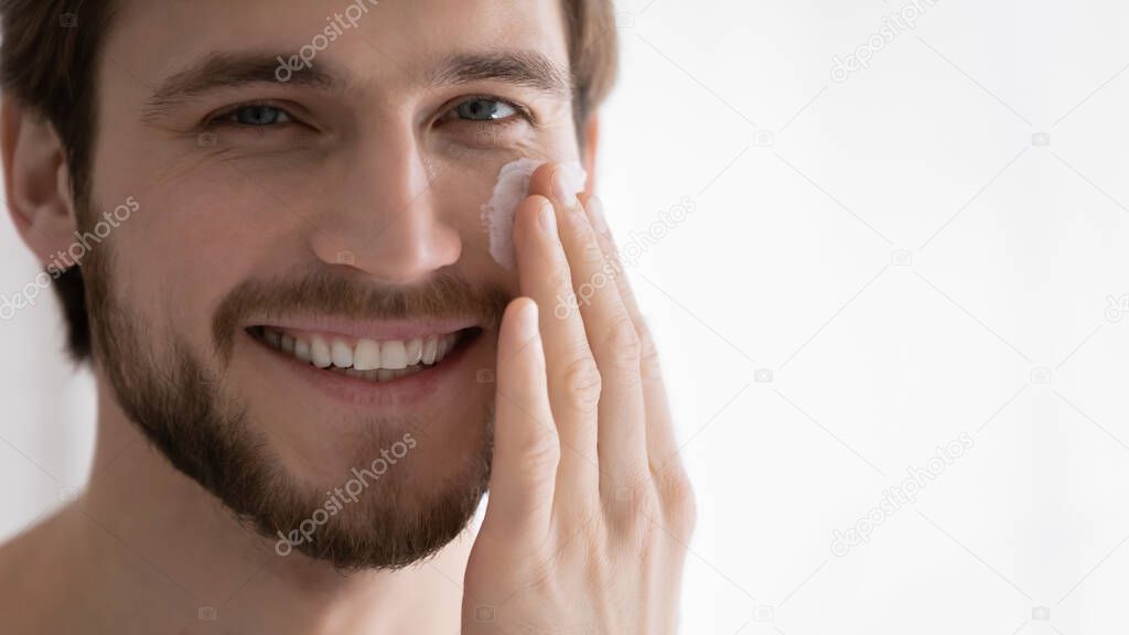 Close up smiling young man applying moisturizing face cream