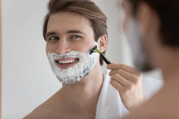 Cerca de reflexión espejo sonriente hombre afeitándose, utilizando la maquinilla de afeitar —  Fotos de Stock