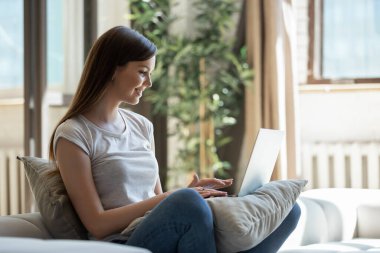 Mutlu gülümseyen genç bayan dizüstü bilgisayarla koltukta oturuyor.