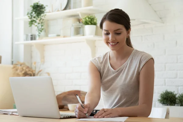 Mujer sonriente trabajando con portátil y tomando notas en la cocina —  Fotos de Stock