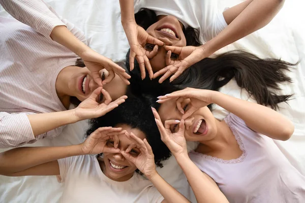 Funny diverse girls lying in bed making binoculars with fingers — Stock Photo, Image