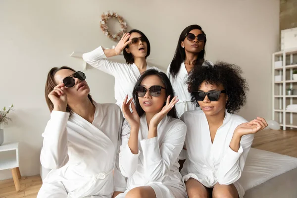 Donne multietniche in accappatoi bianchi e occhiali da sole che si divertono al chiuso — Foto Stock
