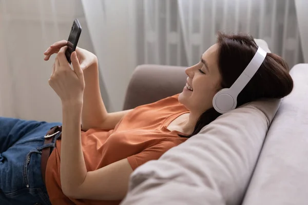 Happy girl in headphones relax on couch using smartphone — Stock Photo, Image