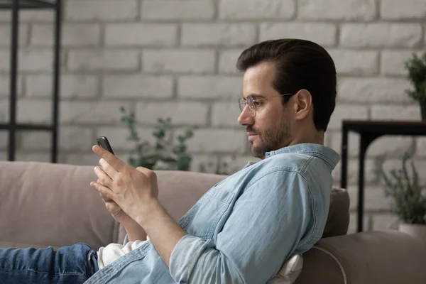 Jovem caucasiano homem relaxar no sofá usando gadget smartphone — Fotografia de Stock