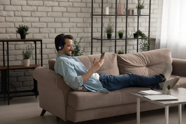 Kulaklıklı mutlu beyaz adam evde rahatlarken akıllı telefon kullanıyor. — Stok fotoğraf