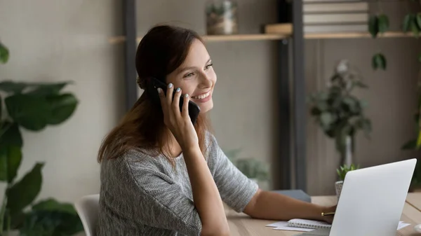 Smiling woman talk on smartphone working on laptop