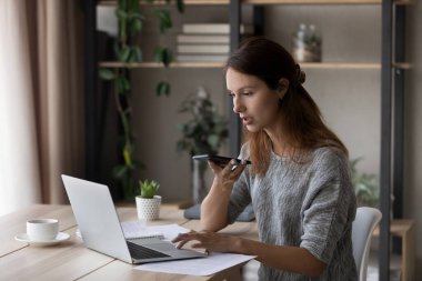 Genç beyaz kadın dizüstü bilgisayarda çalışıyor ve cep telefonuyla konuşuyor.