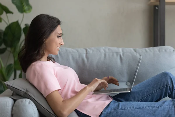 Giovane donna etnica rilassarsi sul divano utilizzando il computer portatile — Foto Stock