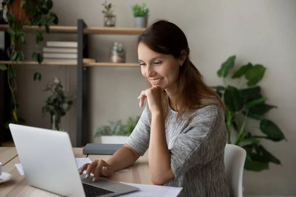 Gülümseyen genç kadın, masaya otur ve dizüstü bilgisayara bak. — Stok fotoğraf