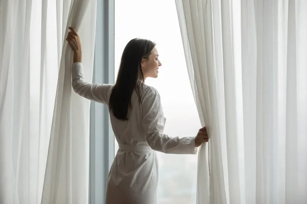 Sorridente giovane donna che indossa accappatoio tende di apertura in camera da letto — Foto Stock