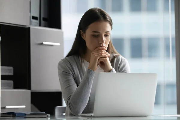 Задумчивая целеустремленная молодая деловая женщина смотрит на экран ноутбука в офисе — стоковое фото