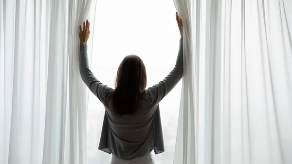 Vista trasera joven mujer abriendo cortinas en la mañana después de despertar —  Fotos de Stock