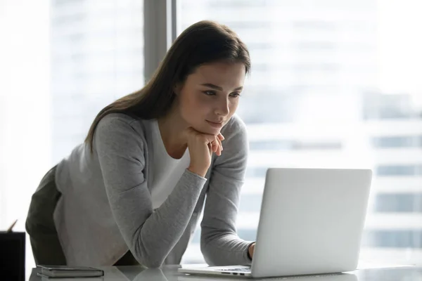 Сосредоточенная молодая деловая женщина, стоящая за столом в офисе, используя ноутбук — стоковое фото