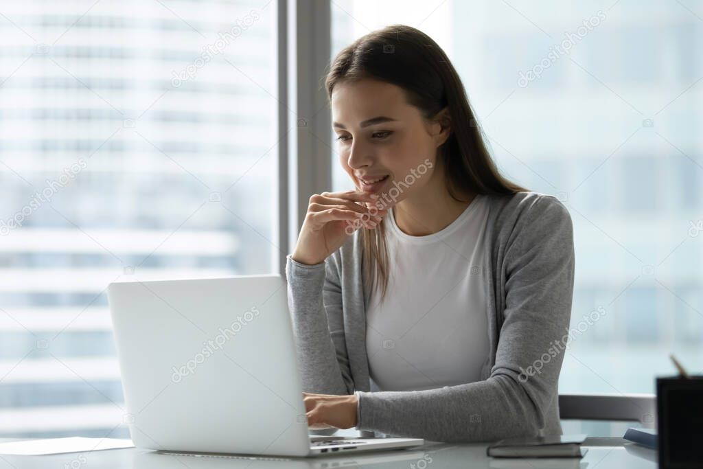Smiling young woman using laptop, sitting at desk in modern office