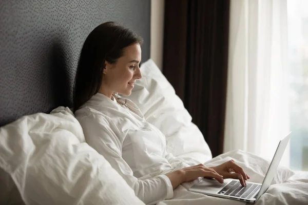 Mujer joven sonriente usando albornoz usando computadora portátil, sentada en la cama — Foto de Stock